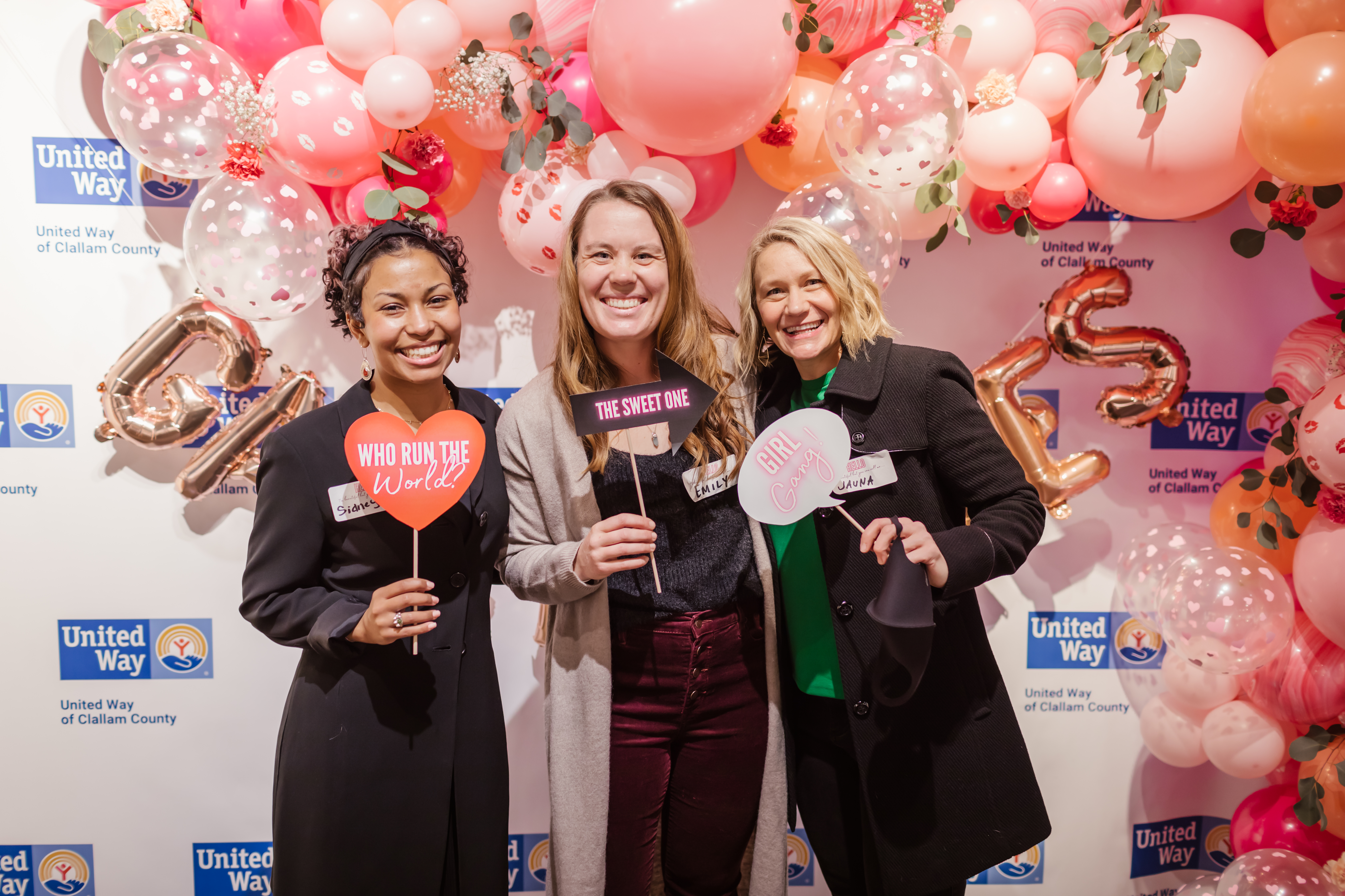 Three smiling women in front of a United Way backdrop with pink and white balloons