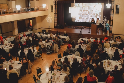 Large room full of people at round tables facing two women presenting on a stage