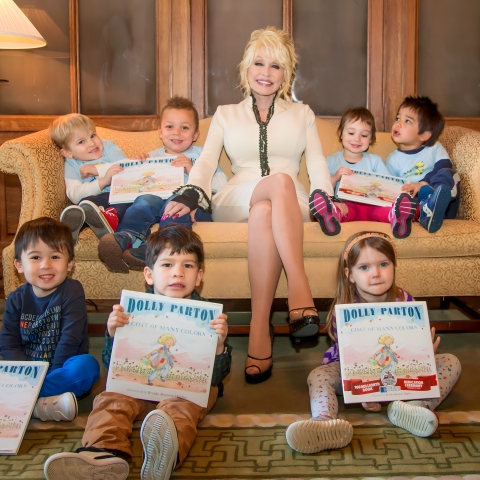 Dolly Parton sitting on a couch surrounded by children holding her book Coat of Many Colors