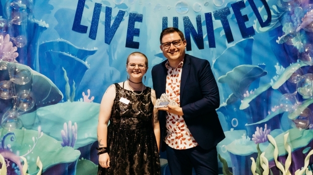 Two people standing in front of an under the sea backdrop. One person holds an award. 